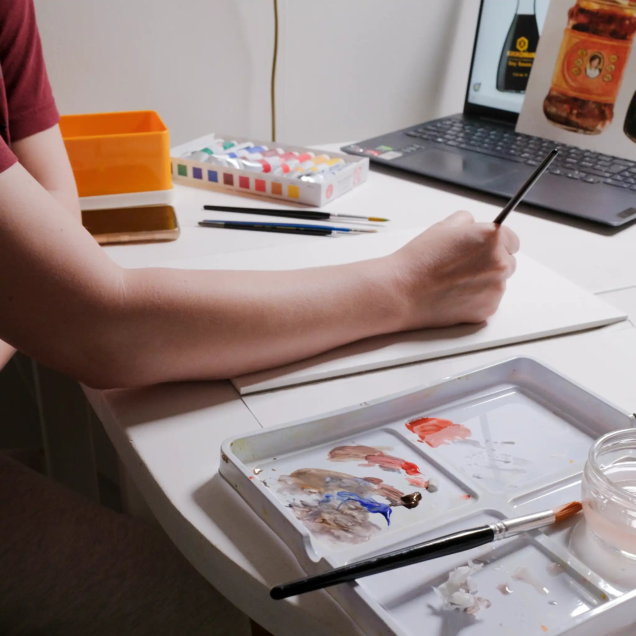 Person painting with watercolors on a table next to a laptop displaying a product image.