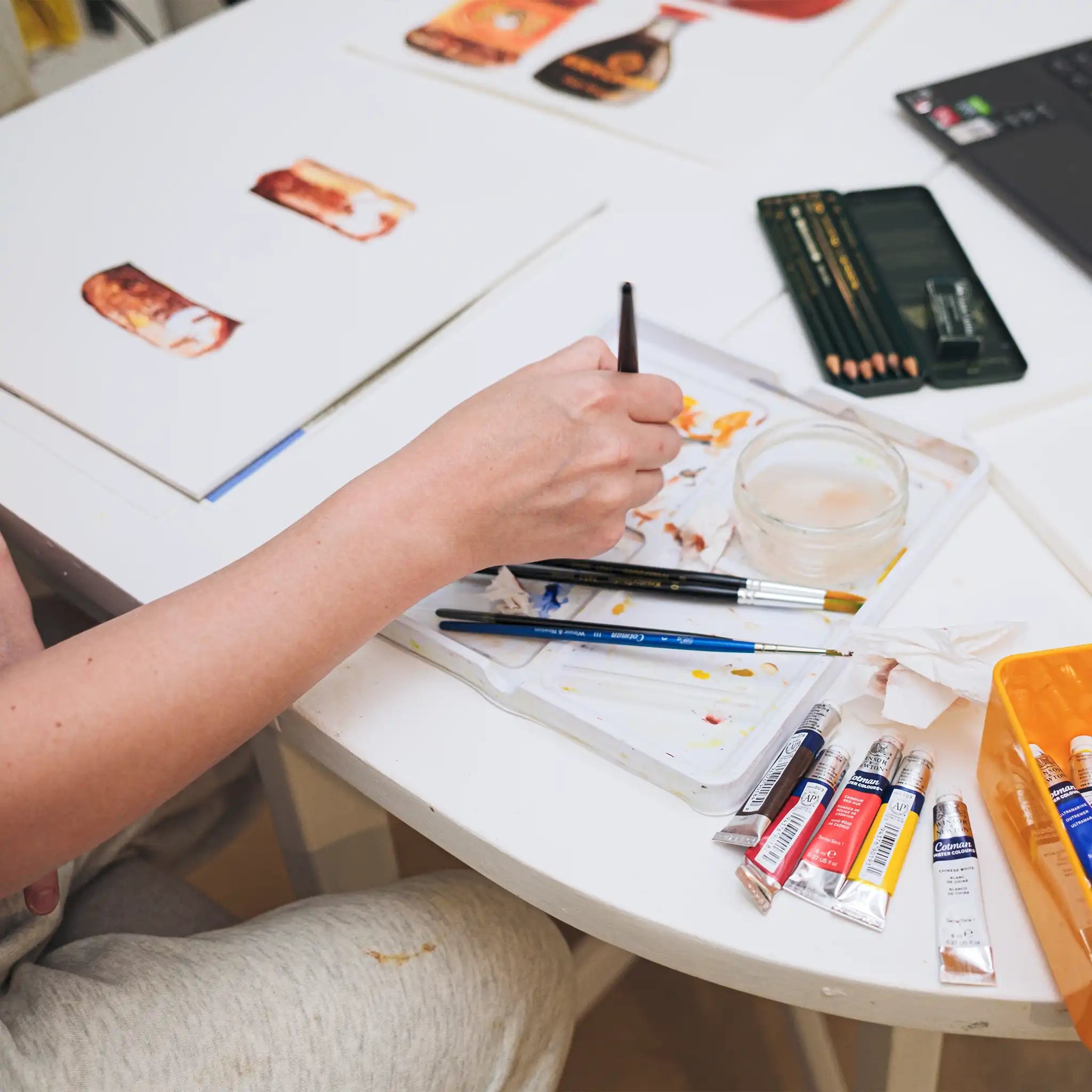 Person painting with watercolors on a white table with art supplies.