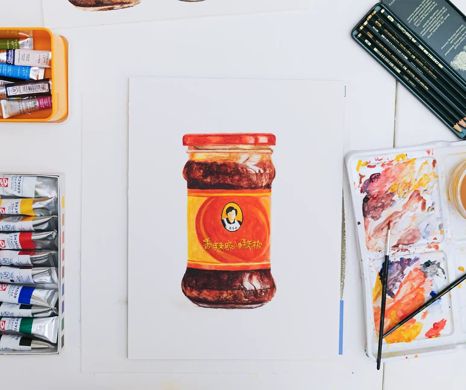 Jar of chili paste on a white surface with art supplies including paint tubes and brushes.