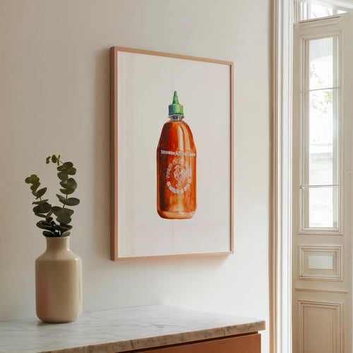 Framed artwork of a sriracha bottle on a wall next to a vase with a plant