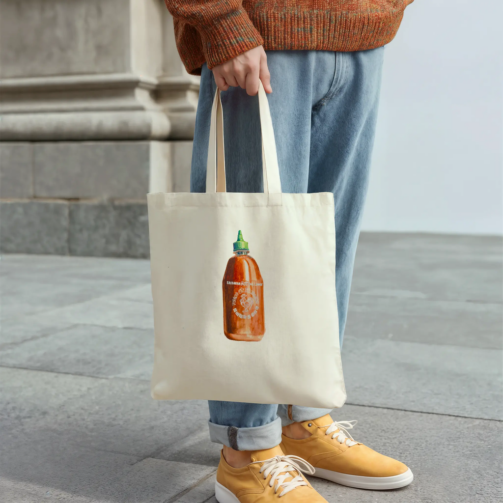 Person holding a tote bag with a sriracha  bottle design, standing on a stone pavement.