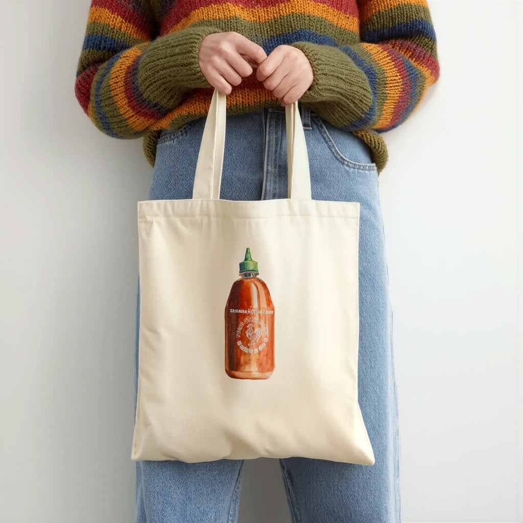 Person holding a tote bag with a sriracha  bottle design, wearing a colorful sweater and jeans.