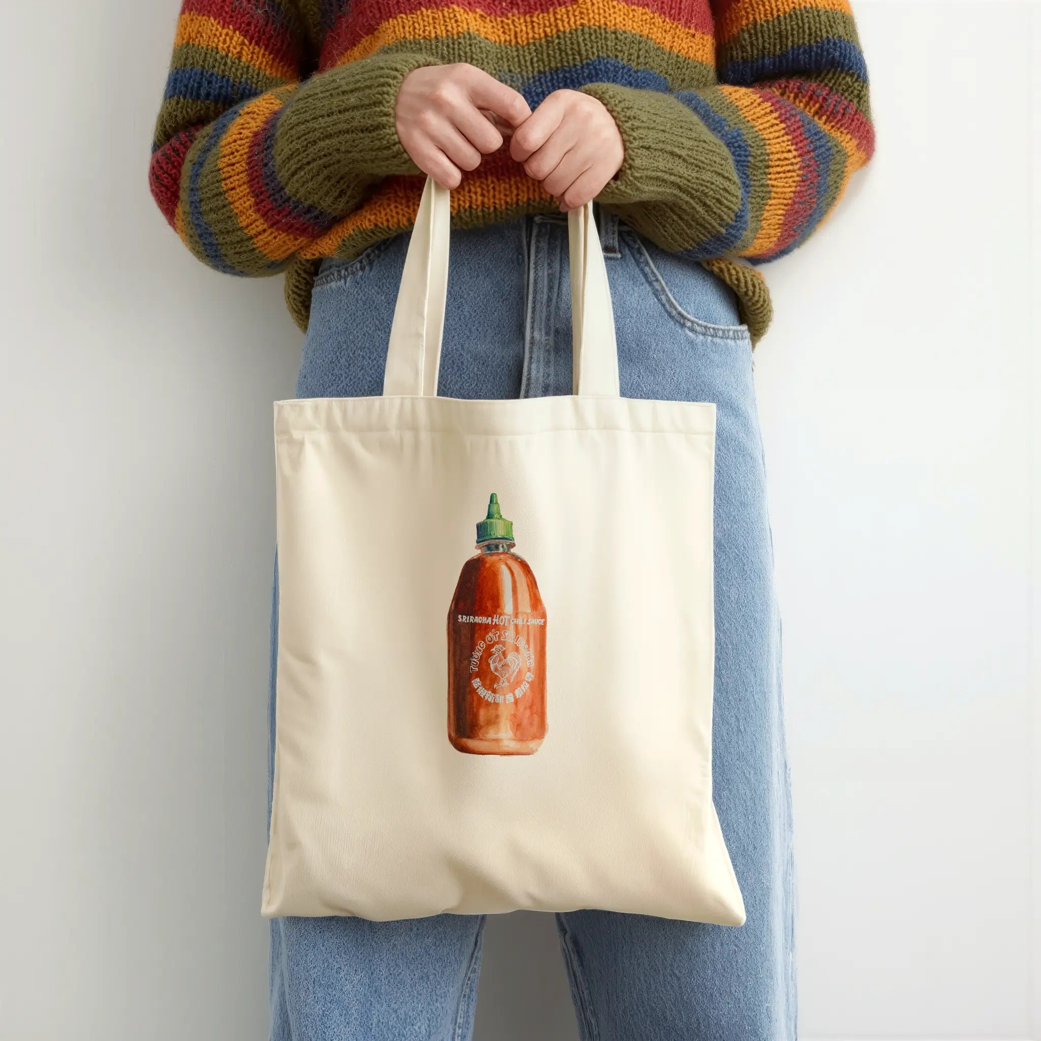 Person holding a tote bag with a sriracha  bottle design, wearing a colorful sweater and jeans.